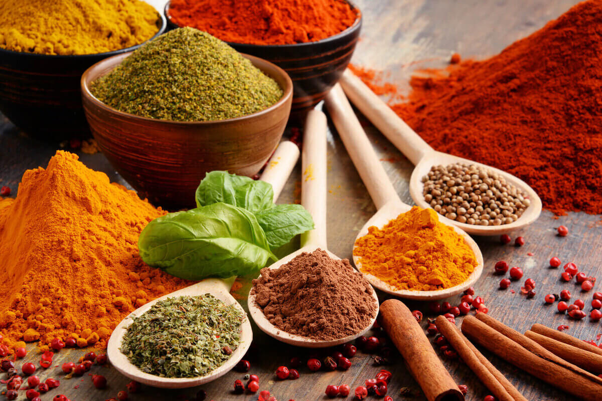 An assortment of colorful spices displayed in wooden spoons and bowls, including turmeric, cinnamon sticks, and ground herbs, with a fresh basil leaf for garnish.