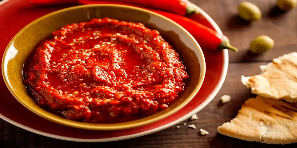 A bowl of vibrant red harissa, a spicy chili pepper paste, with pita bread and olives.