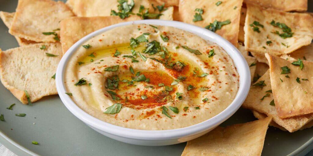 A bowl of creamy hummus garnished with paprika and herbs, surrounded by pita bread chips.