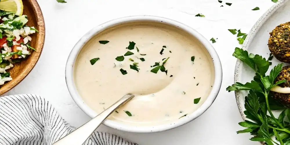 A bowl of creamy tahini sauce sprinkled with parsley. A spoon rests in the bowl.