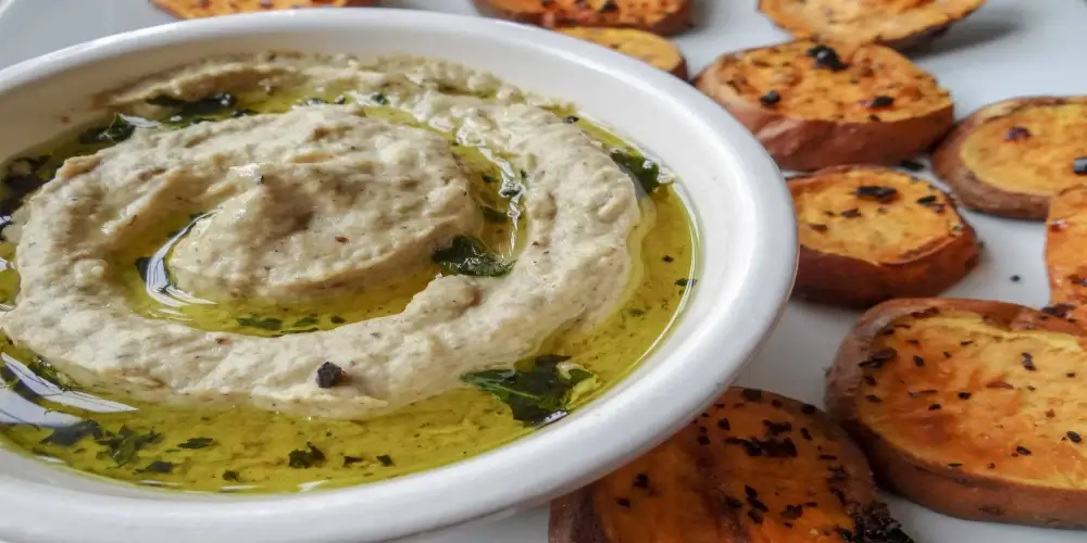 A white bowl filled with creamy baba ghanoush topped with a drizzle of olive oil and garnished with herbs.